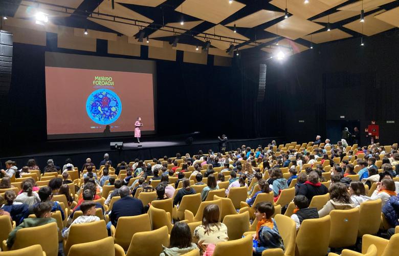 Apertura de MenudoForoAcui. Auditorio cheo de crianzas, coa presentadora no escenario e a imaxe do proxecto na pantalla do fondo.